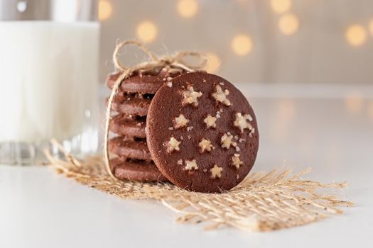 on the table is a stack of round chocolate Christmas cookies with white sugar stars and salt tied with rope, glass of milk, shining garlands in the background