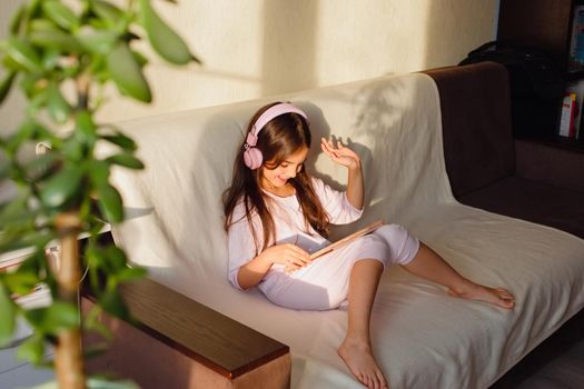 A little beautiful girl in pink home clothes and pink headphones sits on the couch, communicates online using a digital tablet.
