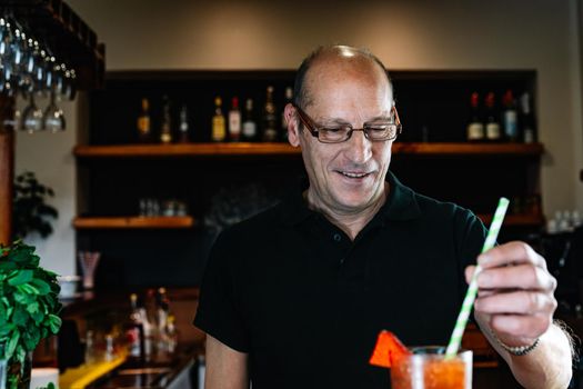 experienced waiter, concentrated and precise, dressed in company uniform, black polo shirt, stirring a cocktail in a crystal glass on the counter of the nightclub. Preparing cocktails for customers. Warm atmosphere and dim light. Horizontal