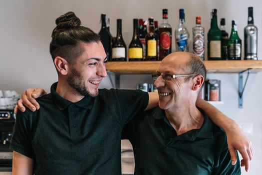 Portrait of two waiters. Mature, experienced and smiling waiter, bald, wearing black polo shirt and glasses. Young waiter with long hair tied back and smiling, wearing black polo shirt. Smiling partners of a small company standing together in their cafeteria. Coworkers looking at each other. Co-workers hugging each other. Warm and pleasant atmosphere, natural light.