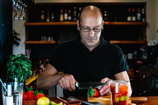 bald adult male, experienced, concentrated and hard-working waiter, dressed in company uniform, black polo shirt, cutting strawberries for a cocktail at the nightclub counter. Preparing cocktails for the customers. Warm atmosphere and dim light. Horizontal