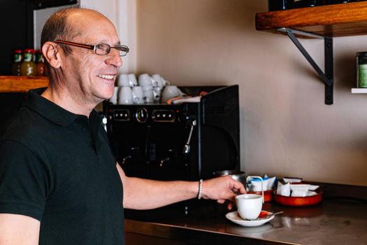 Male waiter, mature, bald, smiling, dressed in company uniform, black polo shirt, Mature waiter preparing a coffee in the coffee machine. Waiter talking to his co-worker. Warm atmosphere and dim lighting. Horizontal