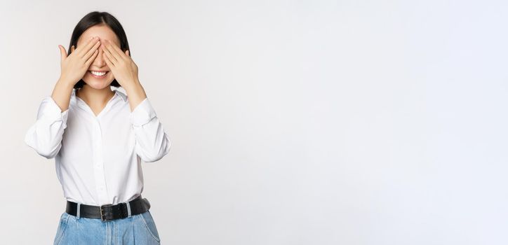 Image of young asian woman close her eyes and smiles, waits for surprise, anticipates, stands over white background.