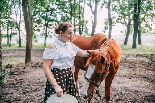 photo of a young smiling blonde, in a white blouse and skirt, with a horse, in a summer forest.