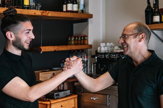 Portrait of two waiters. Mature, experienced and smiling waiter, bald, wearing black polo shirt and glasses. Young waiter with long hair tied back and smiling, wearing black polo shirt. Smiling coworkers high-fiving each other. Coworkers with good relationship. Coworkers hugging each other. Warm and pleasant atmosphere, natural light.