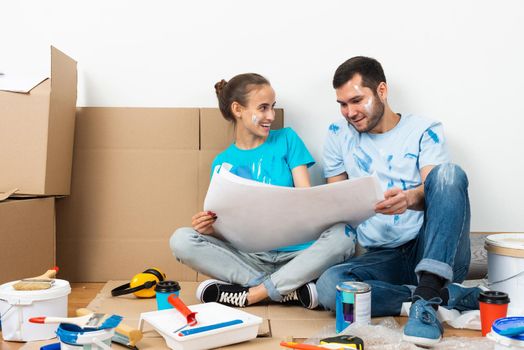 Couple discussing renovating plan of their new home. Smiling young man and woman together sitting on floor among cardboard boxes. House remodeling and interior redesign. People looking at blueprint.