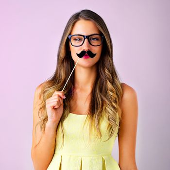 Studio shot of a young woman holding a mustache prop to her face against a pink background.