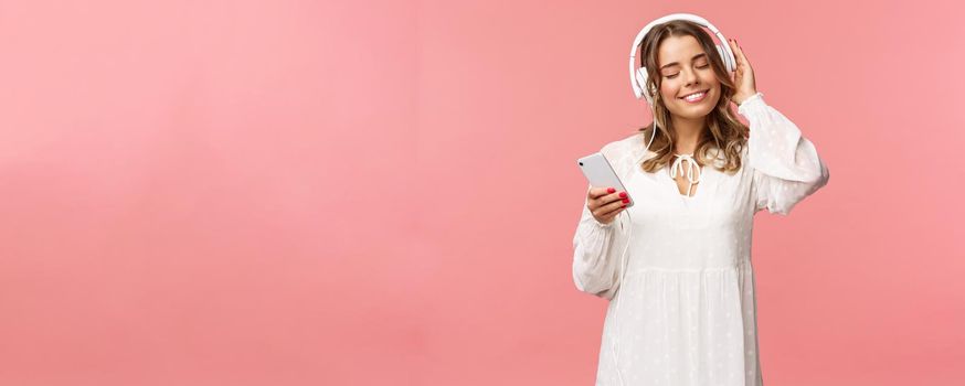 Portrait of tender happy and cute blond girl in white dress, close eyes and smiling pleased as listening music in headphones, feeling warmth and happiness enjoying favorite track, hold mobile phone.