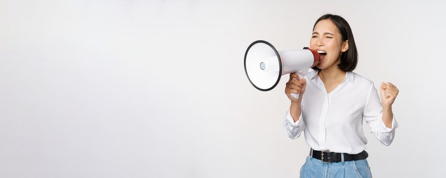 Enthusiastic asian woman, girl activist shouting at protest, using megaphone, looking confident, talking in loudspeaker, protesting, standing over white background.