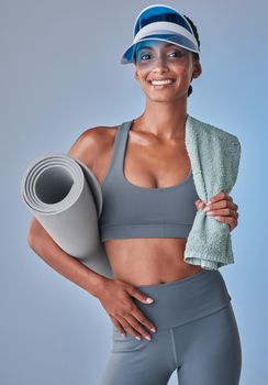 Studio shot of a fit young woman holding a towel and exercise mat against a grey background.