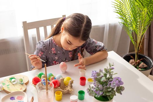 A cute little girl sits at a white table near the window, prepares for the Easter holiday, paints eggs with a brush.