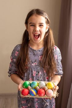 Funny emotional little brunette girl in a cute dress standing in a room with curtains holding a cardboard tray with multi-colored hand-painted eggs