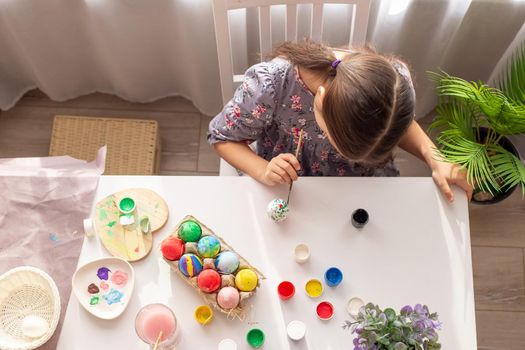 A cute little girl in dress, sits at a white table near the window, paints eggs with a brush. Top view