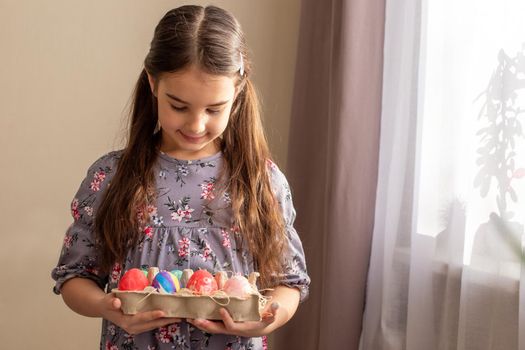 Cute little brunette girl, in cute dress, standing in a room near a window with curtains, holding a cardboard tray with multi-colored hand-painted eggs, looking at them