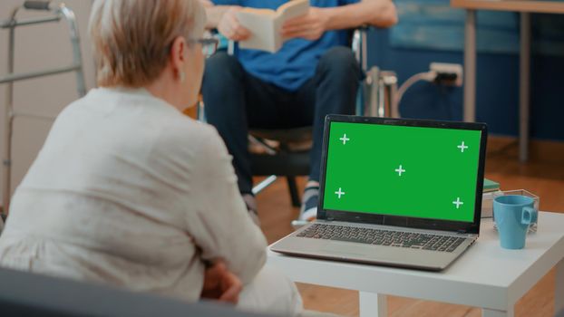 Old woman with disability analyzing green screen on computer. Elder person with crutches looking at chroma key template with blank mock up background and isolated copy space on laptop.