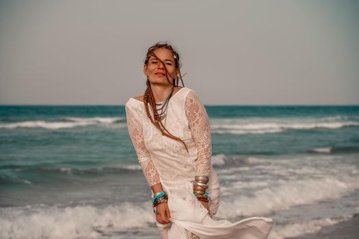 Model in boho style in a white long dress and silver jewelry on the beach. Her hair is braided, and there are many bracelets on her arms