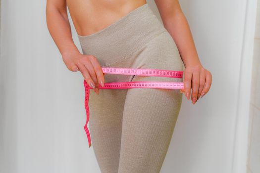 Cropped view of slim woman measuring hips with tape measure at home, close up. An unrecognizable European woman checks the result of a weight loss diet or liposuction indoors. Healthy lifestyle