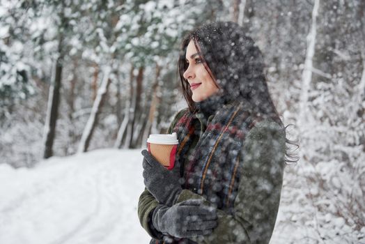 Warm drink helping keeps the heat. Girl in warm clothes with cup of coffee have a walk in the winter forest.