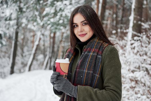 Beautiful brunette. Girl in warm clothes with cup of coffee have a walk in the winter forest.