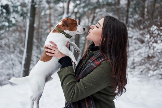 Giving a lovely kiss. Smiling brunette having fun while walking with her dog in the winter park.