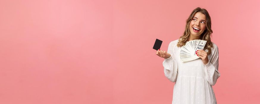 Portrait of pleased attractive blond girl in white dress, daydreaming about what buy, shopping with girlfriends, holding money and credit card, use cash, standing pink background.
