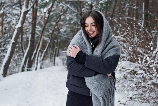 Warming with some good clothes. Portrait of charming woman in the black jacket and grey scarf in the winter forest.