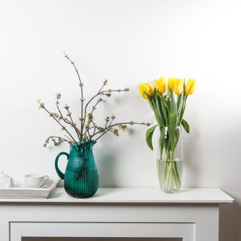 Bouquet of yellow tulip in the glass vase and American maple branches in green glass jar on white table