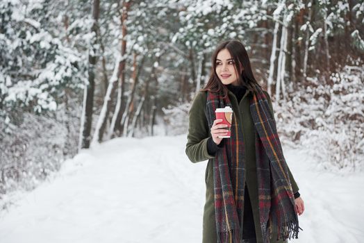 Admires nature. Girl in warm clothes with cup of coffee have a walk in the winter forest.