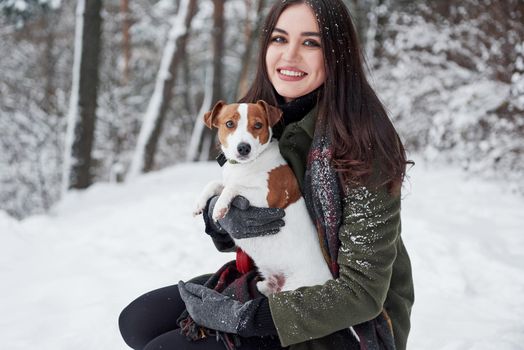 Holds pet in the hands. Smiling brunette having fun while walking with her dog in the winter park.