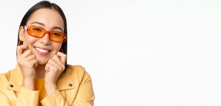 Close up portrait of trendy asian woman in sunglasses, touching her face, looking romantic, smiling at camera, standing over white background.