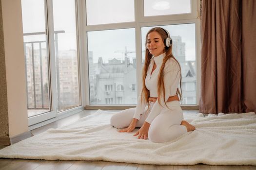 Side view portrait of relaxed woman listening to music with headphones lying on carpet at home. She is dressed in a white tracksuit