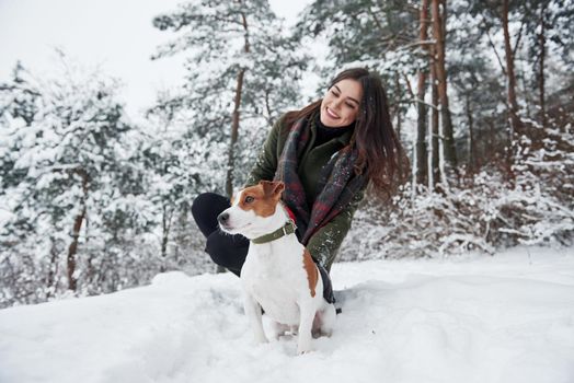 Sitting together. Smiling brunette having fun while walking with her dog in the winter park.