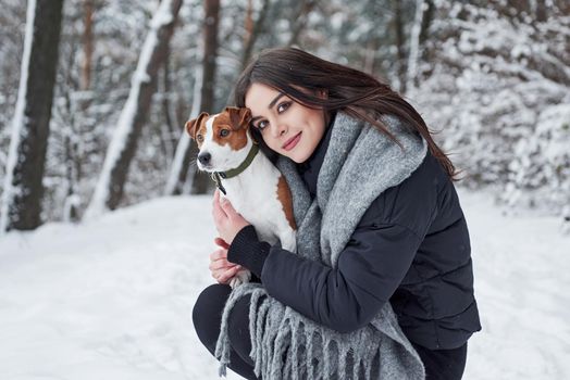 Girl looking to the camera, pet to the side. Smiling brunette having fun while walking with her dog in the winter park.