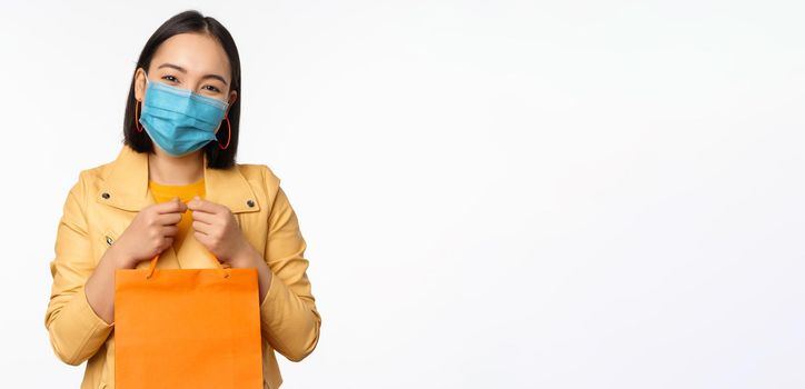 Covid-19 and shop concept. Young asian stylish woman, wearing medical face mask, holding shopping bag, going in malls during pandemic, white background.