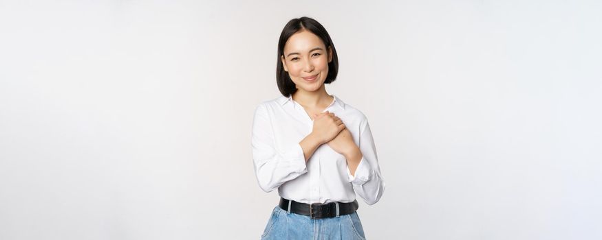 Image of caring young beautiful asian woman, holding hands on heart, looking pleased and happy, smiling at camera, standing over white background.