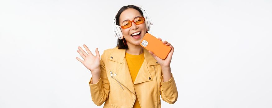 Happy stylish asian woman in sunglasses, listening music in headphones, holding smartphone, singing and dancing, standing over white background.