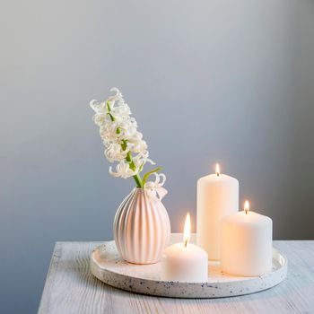 A bouquet of white cut hyacinth a in a small white corrugated vase and three large burning candles on a round tray are on a beige table. Place for text