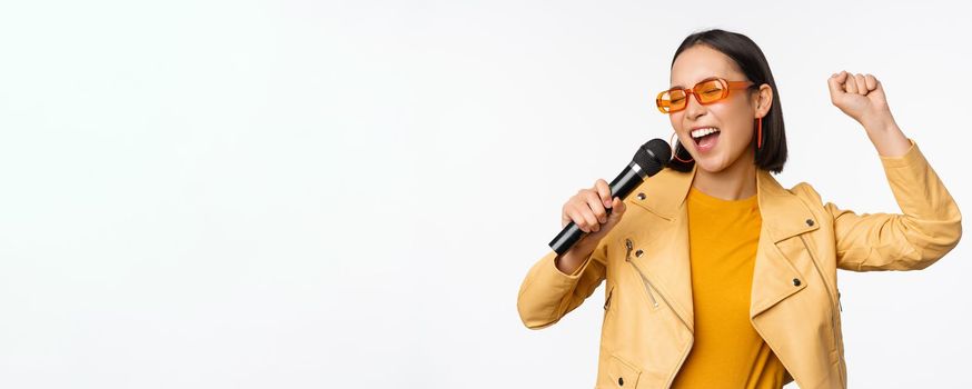 Stylish asian girl in sunglasses, singing songs with microphone, holding mic and dancing at karaoke, posing against white background.