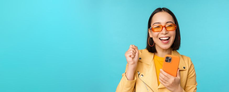 Happy stylish asian girl using smartphone and laughing, smiling at camera, standing over blue background. Copy space