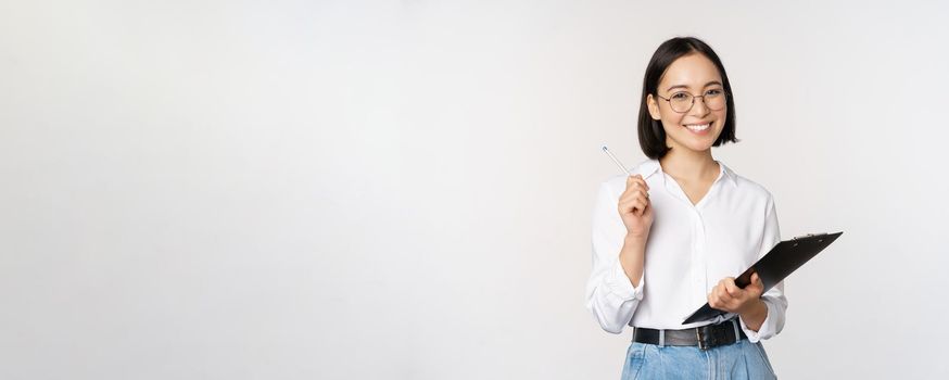 Smiling young asian woman taking notes with pen on clipboard, looking happy, standing against white background.