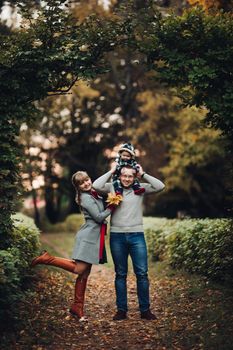 Portrait of attractive young mother and handsome smiling father wearing glasses holding their beautiful lovely baby girl on hands standing against green hedge in autumnal park. They are smiling and looking at camera.