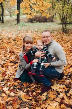 Portrait of attractive young mother and handsome smiling father wearing glasses holding their beautiful lovely baby girl on hands standing against green hedge in autumnal park. They are smiling and looking at camera.