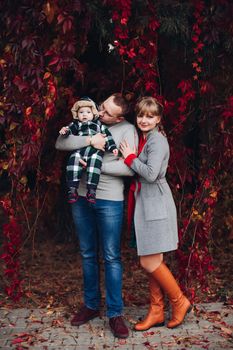 Portrait of attractive young mother and handsome smiling father wearing glasses holding their beautiful lovely baby girl on hands standing against green hedge in autumnal park. They are smiling and looking at camera.