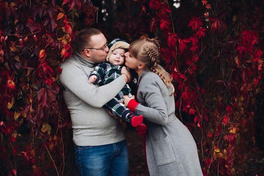 Portrait of attractive young mother and handsome smiling father wearing glasses holding their beautiful lovely baby girl on hands standing against green hedge in autumnal park. They are smiling and looking at camera.