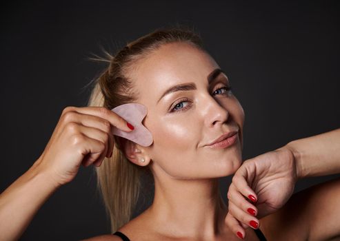 Close-up beautiful European woman doing facial lifting smoothing lymphatic drainage massage on her face, using a gua-sha jade stone roller massager, caring of her skin health and youth