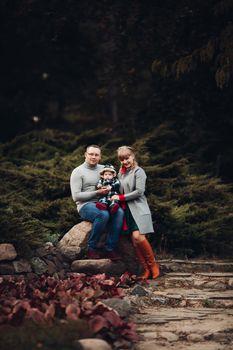 Portrait of attractive young mother and handsome smiling father wearing glasses holding their beautiful lovely baby girl on hands standing against green hedge in autumnal park. They are smiling and looking at camera.