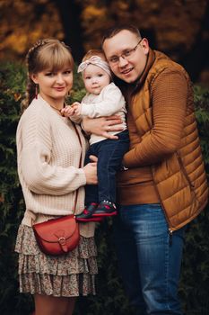 Portrait of attractive young mother and handsome smiling father wearing glasses holding their beautiful lovely baby girl on hands standing against green hedge in autumnal park. They are smiling and looking at camera.