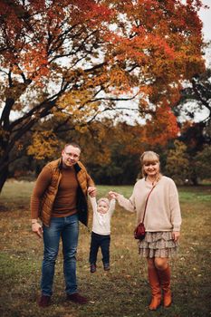 Portrait of attractive young mother and handsome smiling father wearing glasses holding their beautiful lovely baby girl on hands standing against green hedge in autumnal park. They are smiling and looking at camera.