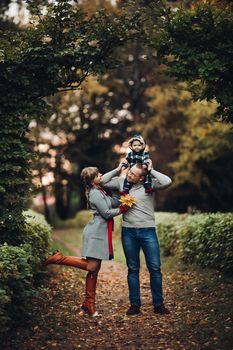 Portrait of attractive young mother and handsome smiling father wearing glasses holding their beautiful lovely baby girl on hands standing against green hedge in autumnal park. They are smiling and looking at camera.