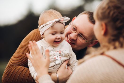 Portrait of attractive young mother and handsome smiling father wearing glasses holding their beautiful lovely baby girl on hands standing against green hedge in autumnal park. They are smiling and looking at camera.